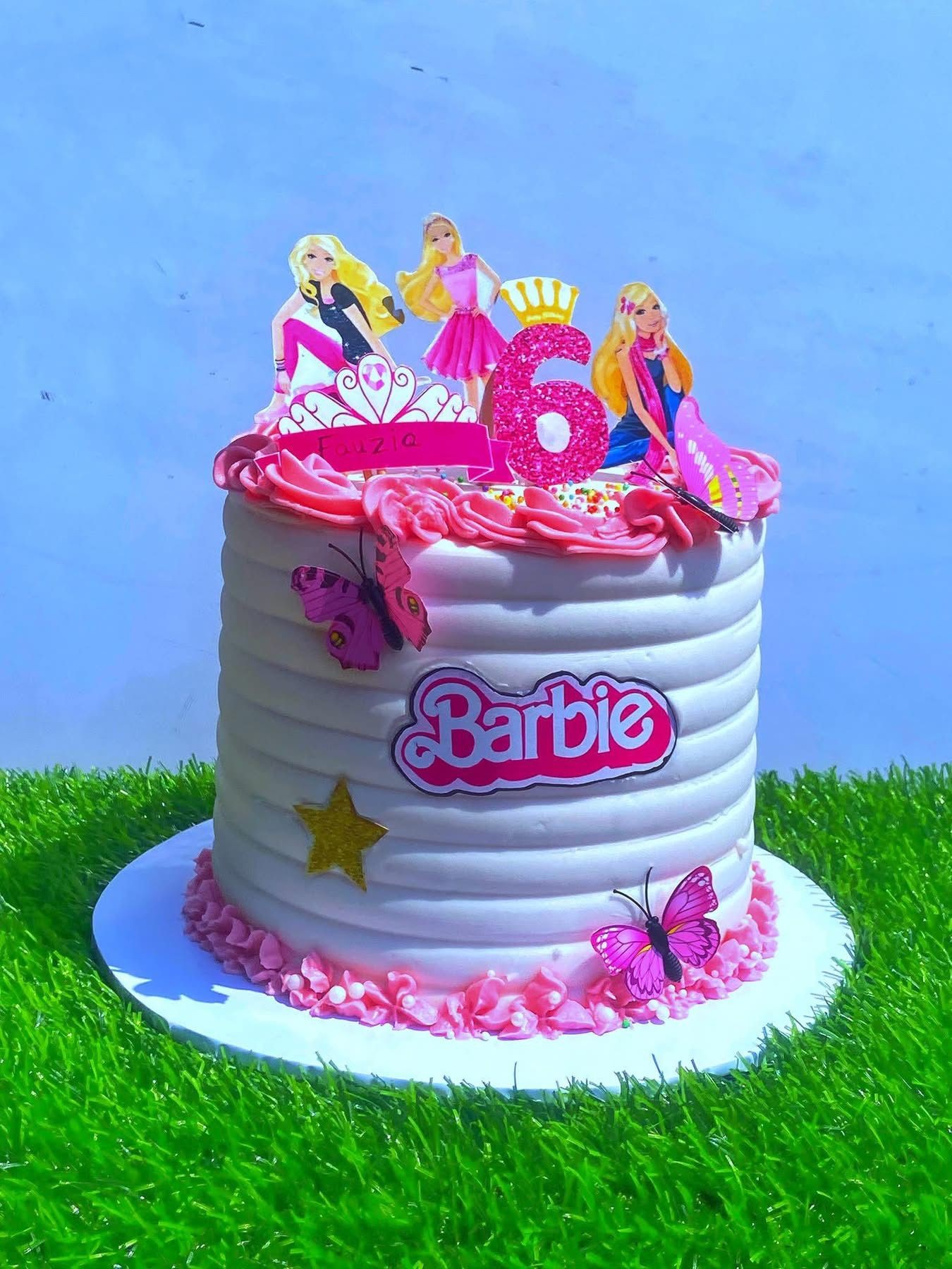 Tall Barbie-themed birthday cake with pink decorations and butterfly toppers on a grassy surface.