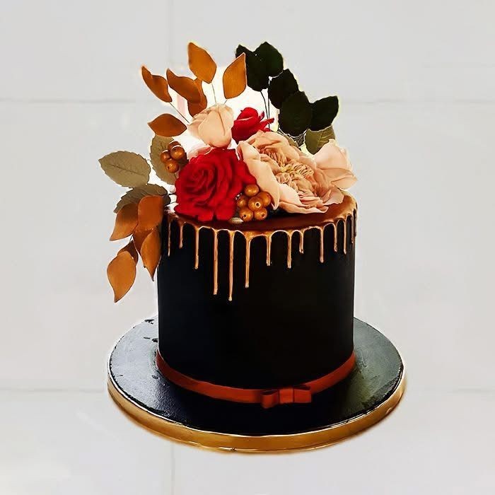 Elegant black cake with gold drip and floral decorations, including red and pink flowers and golden leaves.