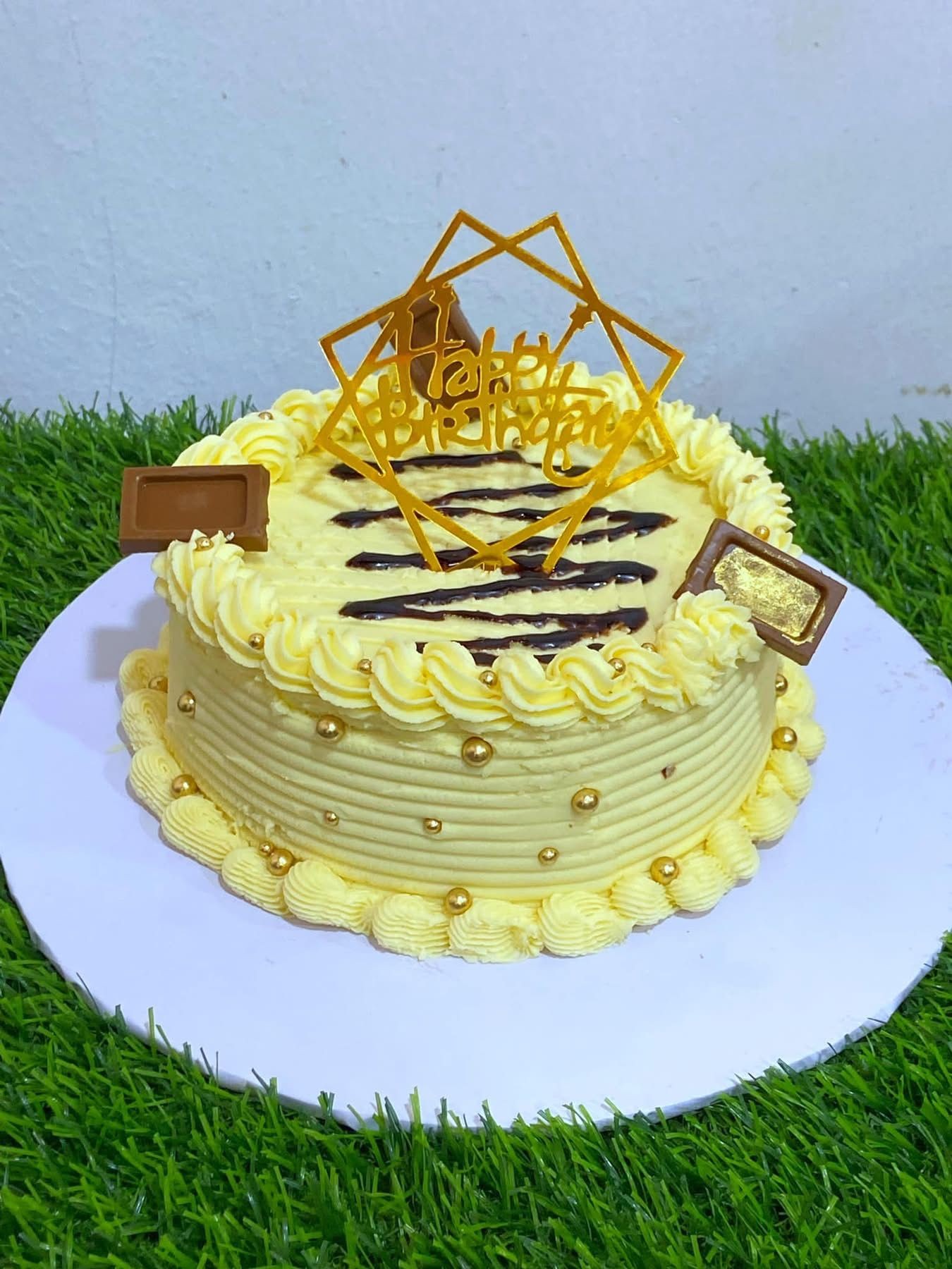 Yellow frosted birthday cake with chocolate decorations and a geometric topper, placed on grass.