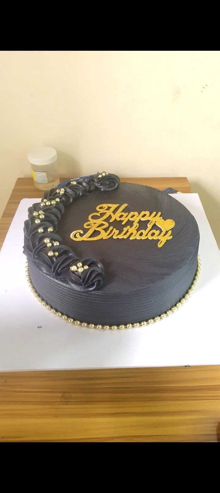 Dark-themed birthday cake with gold lettering and decorative pearls on a wooden table.