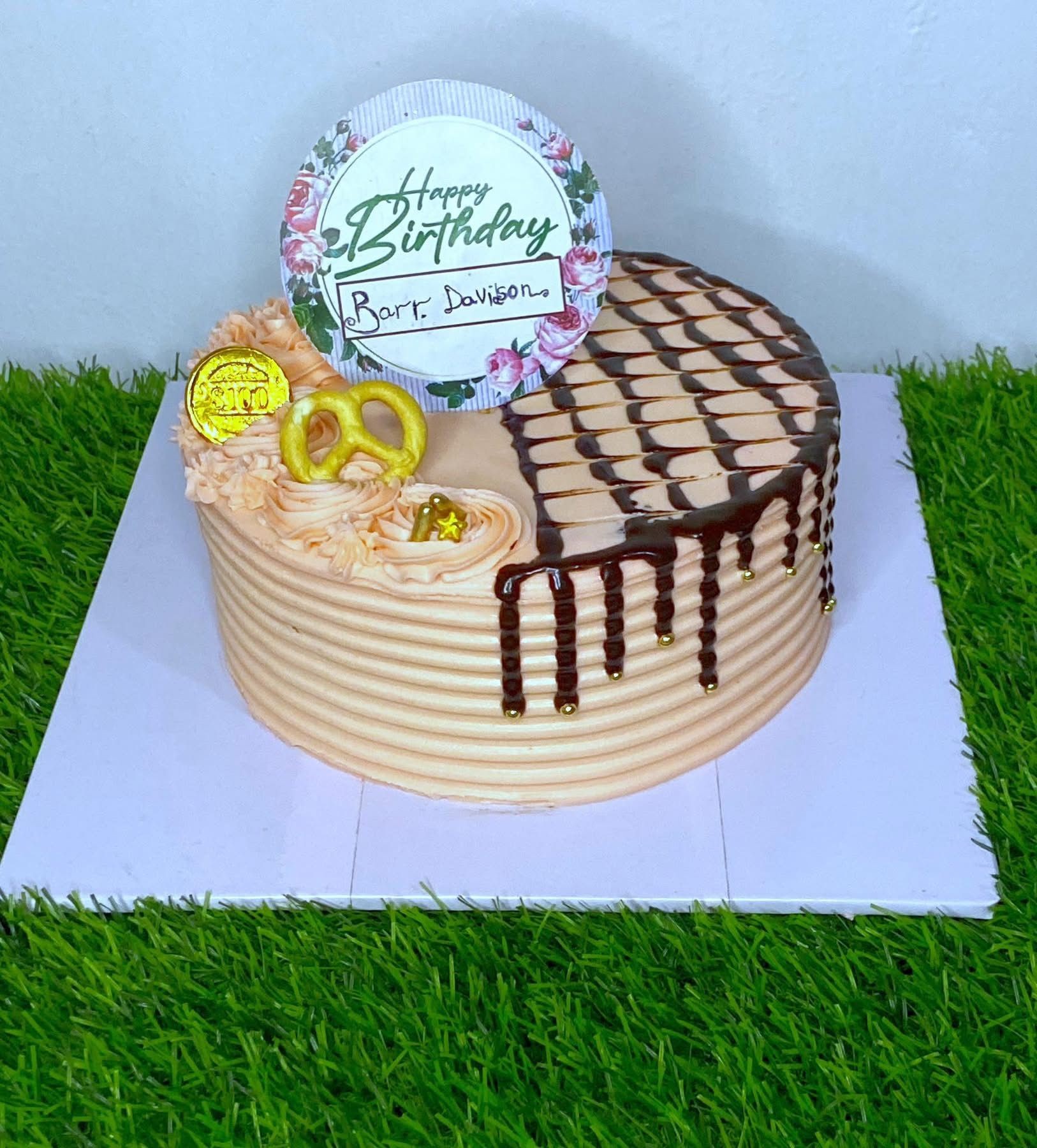 Round birthday cake with chocolate drizzle, decorative pretzel, and floral topper on green surface.