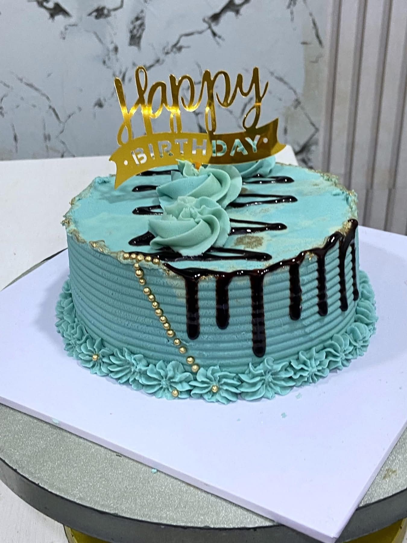 Blue birthday cake with chocolate drips, floral icing, and gold 'Happy Birthday' topper.