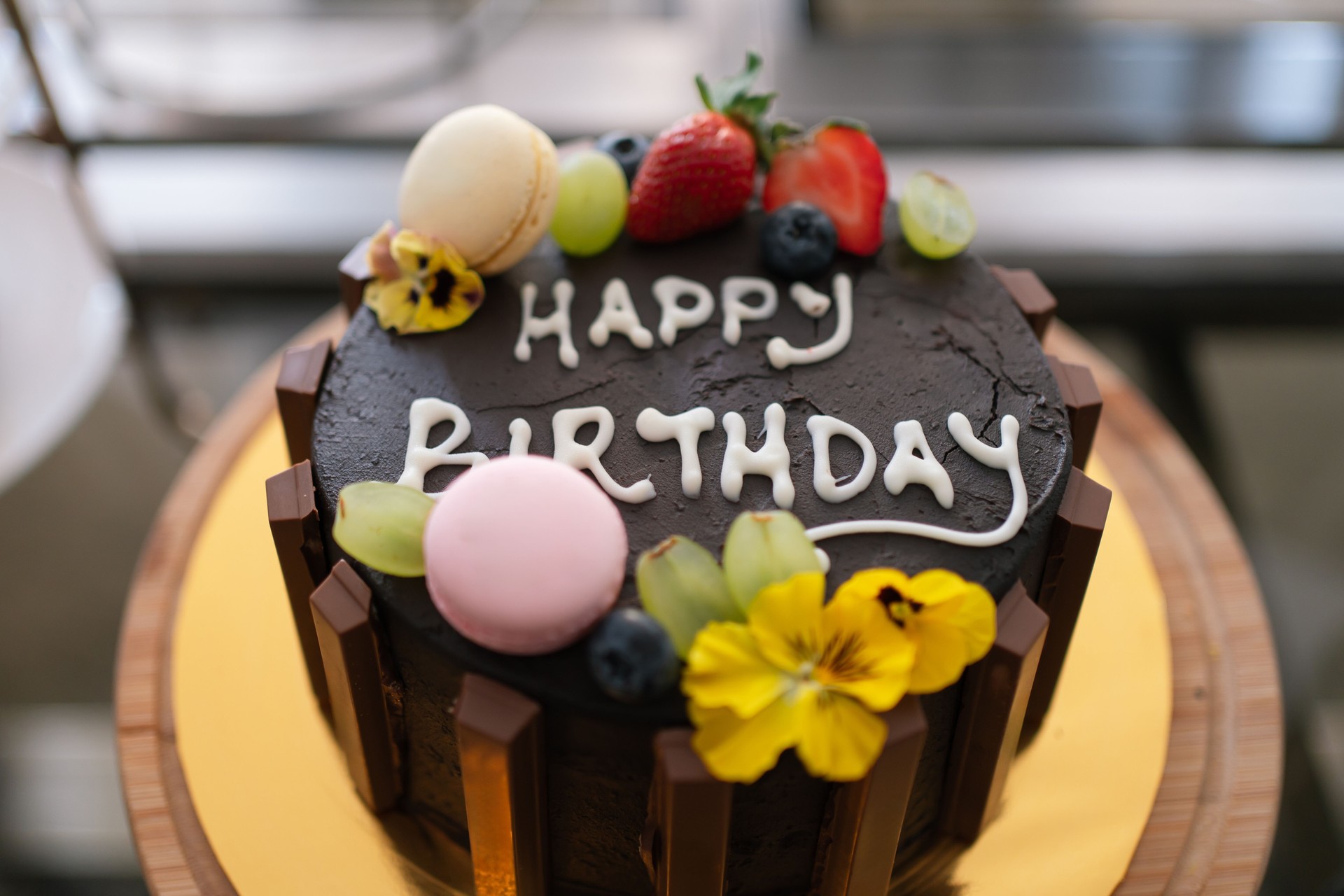 Chocolate cake decorated with edible flowers, fruits, and macarons, featuring handwritten "Happy Birthday".